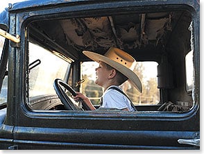 A young boy wearing a cowboy hat smiling behind the wheel of a vintage truck